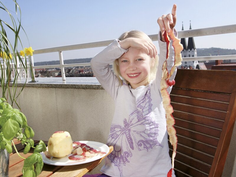 Ein kleines Mädchen sitzt fröhlich auf einem sonnigen Balkon und hält Gemüsereste in den Händen.