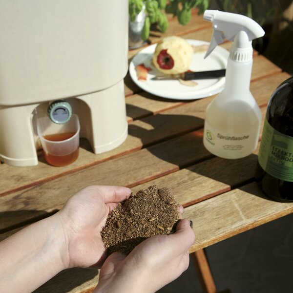 Nahaufnahme von Händen, die fermentiertes Bokashi-Material über einem Holztisch mit Sprühflasche halten.