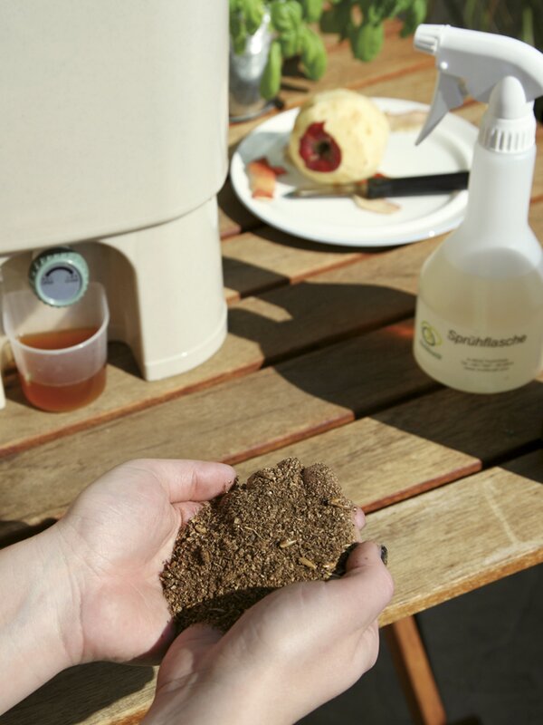 Nahaufnahme von Händen, die fermentiertes Bokashi-Material über einem Holztisch mit Sprühflasche halten.