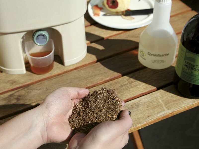 Nahaufnahme von Händen, die fermentiertes Bokashi-Material über einem Holztisch mit Sprühflasche halten.