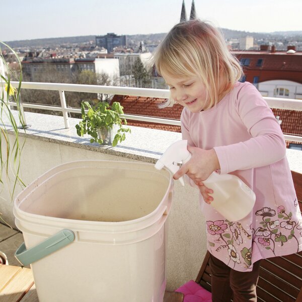 Ein kleines Mädchen nutzt eine Sprühflasche neben einem Bokashi-Eimer auf dem Balkon.