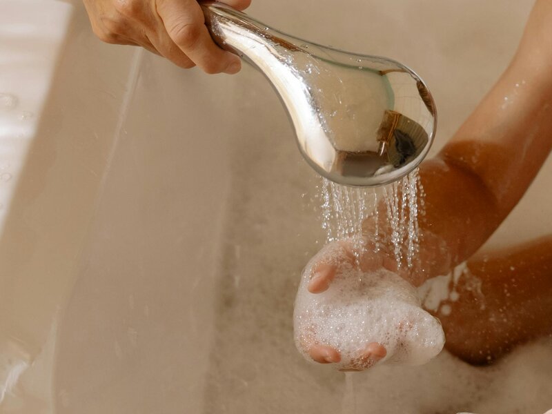 Eine Hand lässt warmes Wasser aus einer Brause in die gefüllte Badewanne laufen.