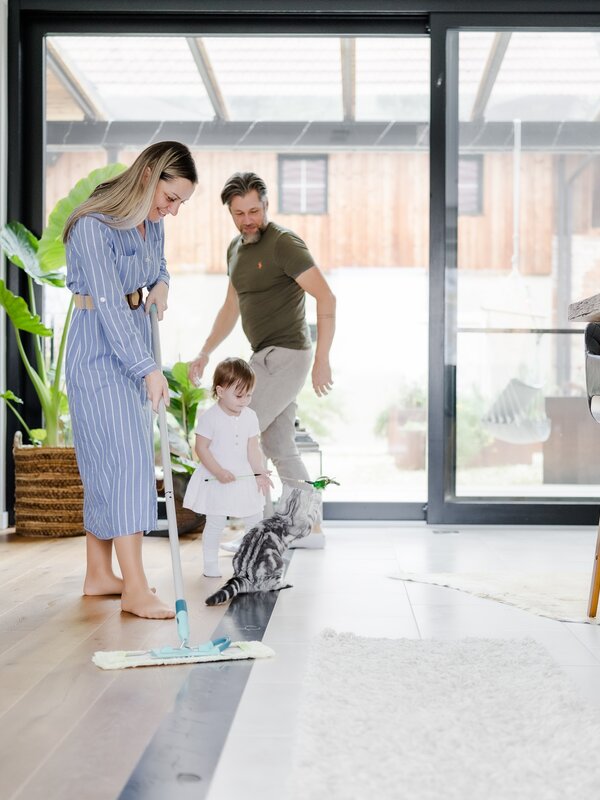 Eine Familie reinigt gemeinsam den Parkettboden in einem sonnendurchfluteten Wohnzimmer.