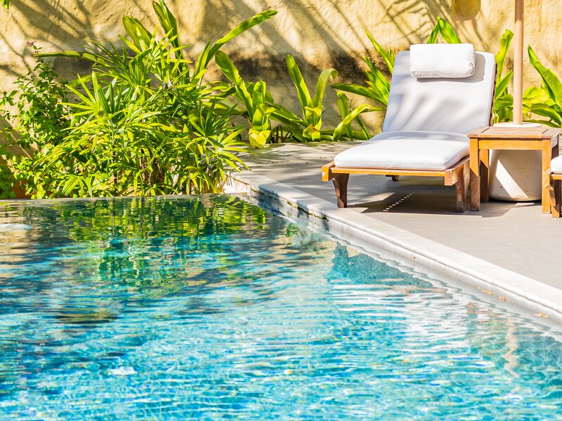 Ein blaues Schwimmbecken im sonnigen Garten mit Sonnenliegen auf der Terrasse.
